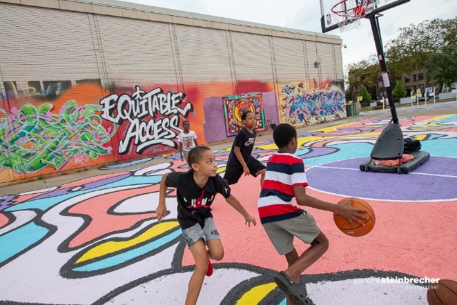 Young people playing basketball on a court covered in lively colors. 'Equitable Access' is written on a wall behind them.