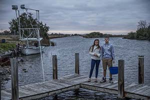 On overcast day, a young woman and man stand at the peak of a dock on edge of calm water with fishing boat with tower in background.