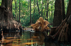 Performer fully covered in costume of gold and red natural materials and talisman objects whirls in wetlands surrounded by giant Cypress trees.