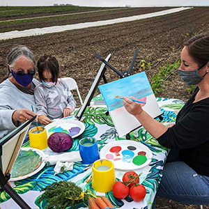 Three generations of women wearing face masks sit at a picnic table in a crop field and paint on small canvases.