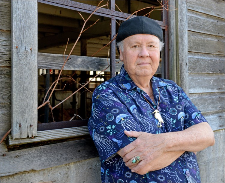 G. Peter Jemison with crossed arms leaning against a window of a wooden structure. He is wearing a blue shirt with a jellyfish design and a black cap.