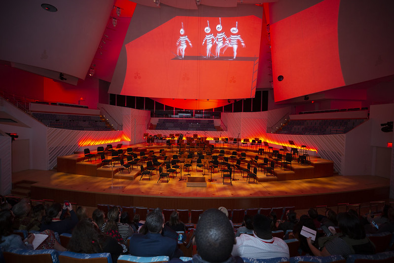 A crowd sits facing a stage set up with chairs for an orchestra. The chairs are empty but above them a video plays. The video casts a red light over the entire room.