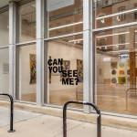 Photo looking through large glass windows into an art gallery. Text on the glass reads: Can you see me?