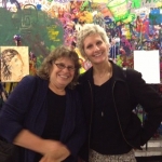 Two smiling people pose in front of a colorful wall of art. They lean toward each other in collegial friendship.