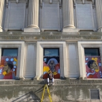 Artist Matthew Cooper sits on a ledge of the old city hall building. Behind him is the center of three portraits of Black women.