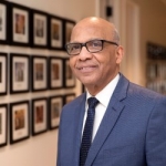 Smiling person wearing glasses and a blue suit in front of a wall with framed art.