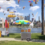 Three-dimensional street vendor carts float in the sky and line a park walkway with a city skyline in the background.