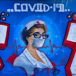 Graffiti style mural painted on a brick wall showing a nurse in traditional white uniform and a white mask with a heartbeat graph behind them and COVID-19 above them. 