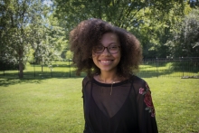 A smiling young Black woman wearing glasses, dark curly hair, and a black top with embroidered floral sleeves.