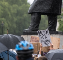 It's a photo of a protest at the foot of a large public art monument. In the foreground a protestor holds up a sign that reads "Racism is a pandemic too." 