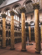 Tall ionic marble columns in the foreground of a photo of a multi-story interior courtyard space with a spouting fountain. Visible in the background are arches lining the walls and a matching set of marble columns.