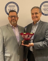 Two people stand in front of a backdrop with the NLGA logo. They both wear gray business jackets and one holds the leadership award, which is a decorative red glass bowl on a small pedestal.