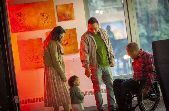 A child interacts with a person in a wheelchair. Two adults are standing next to the child. There are pictures orange, faded paper on the wall behind them.