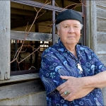 Person leaning against a building with wooden siding, arms crossed, wearing a black hat, blue pattered short-sleeved shirt, necklace with a white bird pendant, and a turquoise ring. 