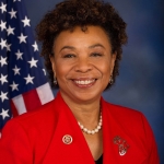 A person sits in front of a blue background and US flag. They have curly dark brown hair and are smiling directly at the camera. They are dressed in a red suit jacket with pins on both lapels.